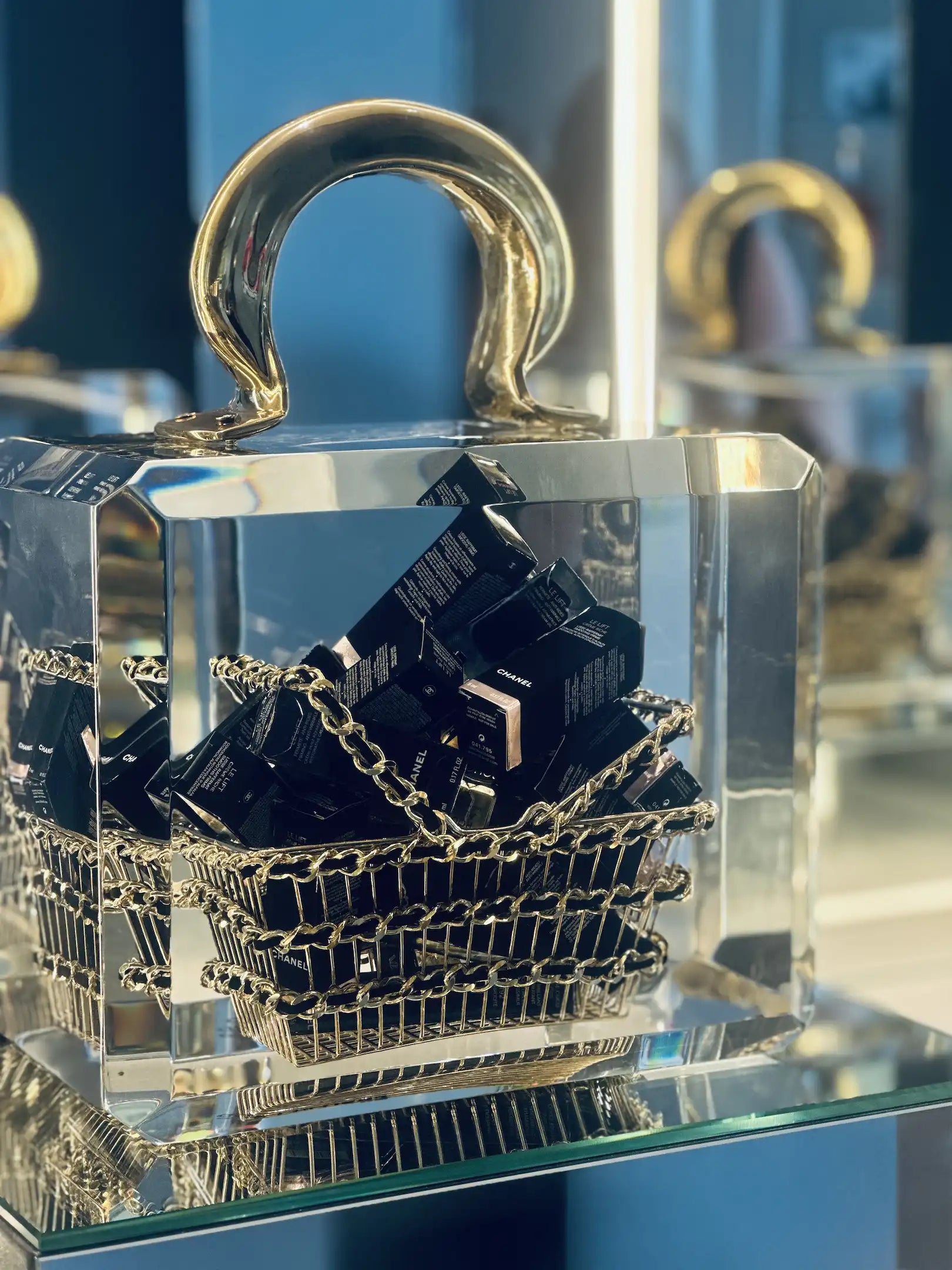 Le sac panier "Chanel overdose" de Fred Allard - une pièce de luxe transparente avec une poignée et une chaîne dorées, remplie de boîtes de cosmétiques Chanel noires - est présenté sur une surface réfléchissante devant un miroir.
