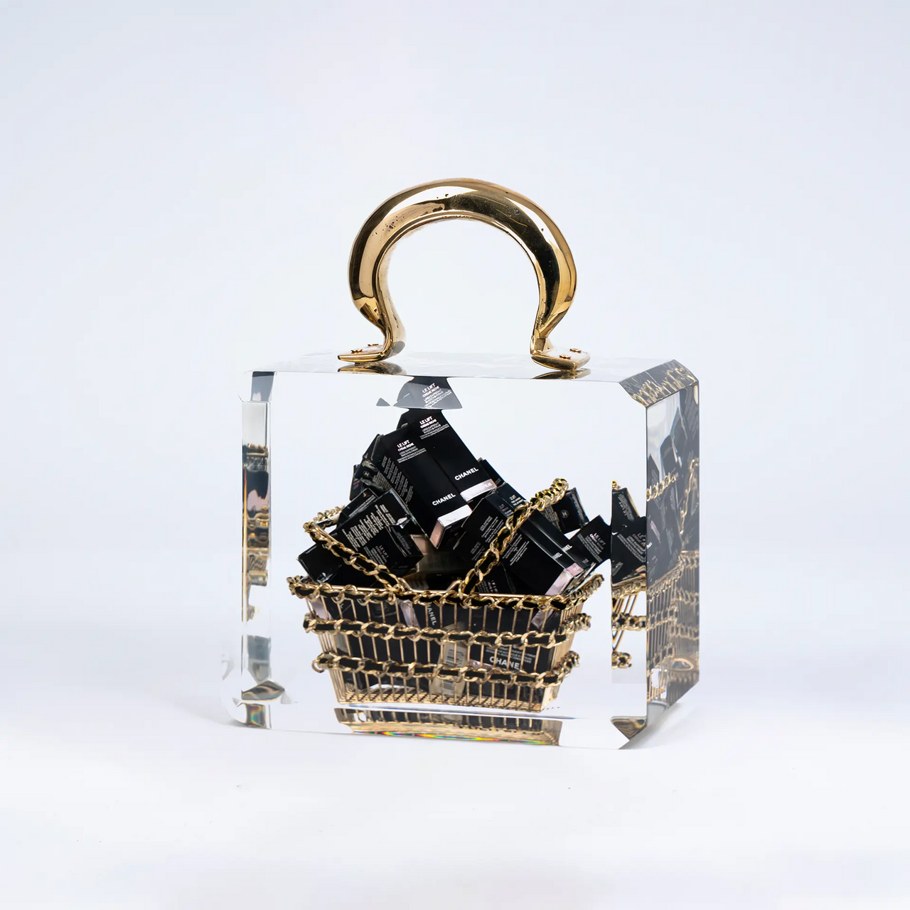 Le Chanel Overdose de Fred Allard est un luxueux basket bag transparent avec une anse dorée, contenant un petit panier doré rempli de boîtes de cosmétiques Chanel noires, le tout sur un fond blanc uni.