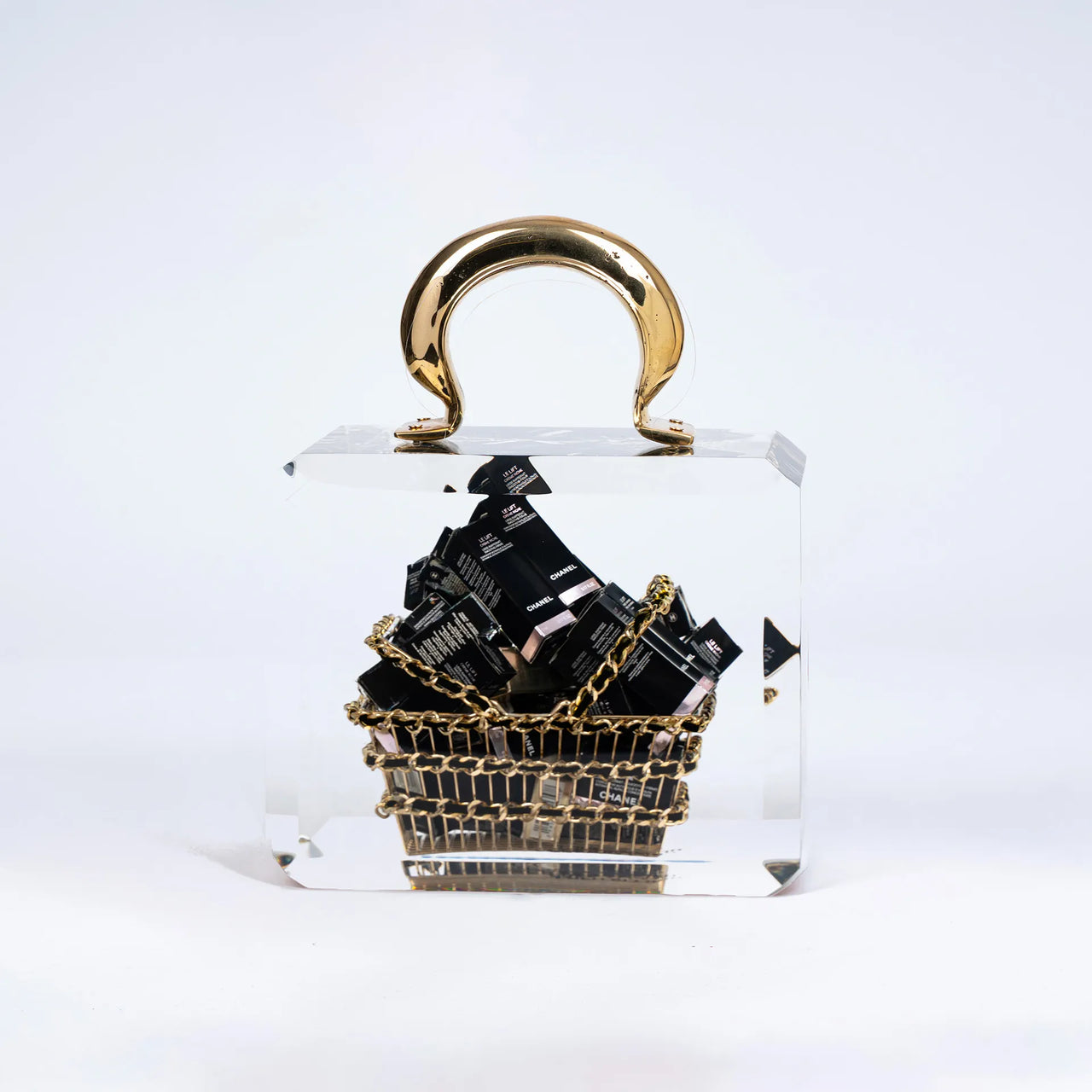 Le Chanel Overdose de Fred Allard est un luxueux basket bag transparent avec une anse dorée, contenant un petit panier doré rempli de boîtes de cosmétiques Chanel noires, le tout sur un fond blanc uni.