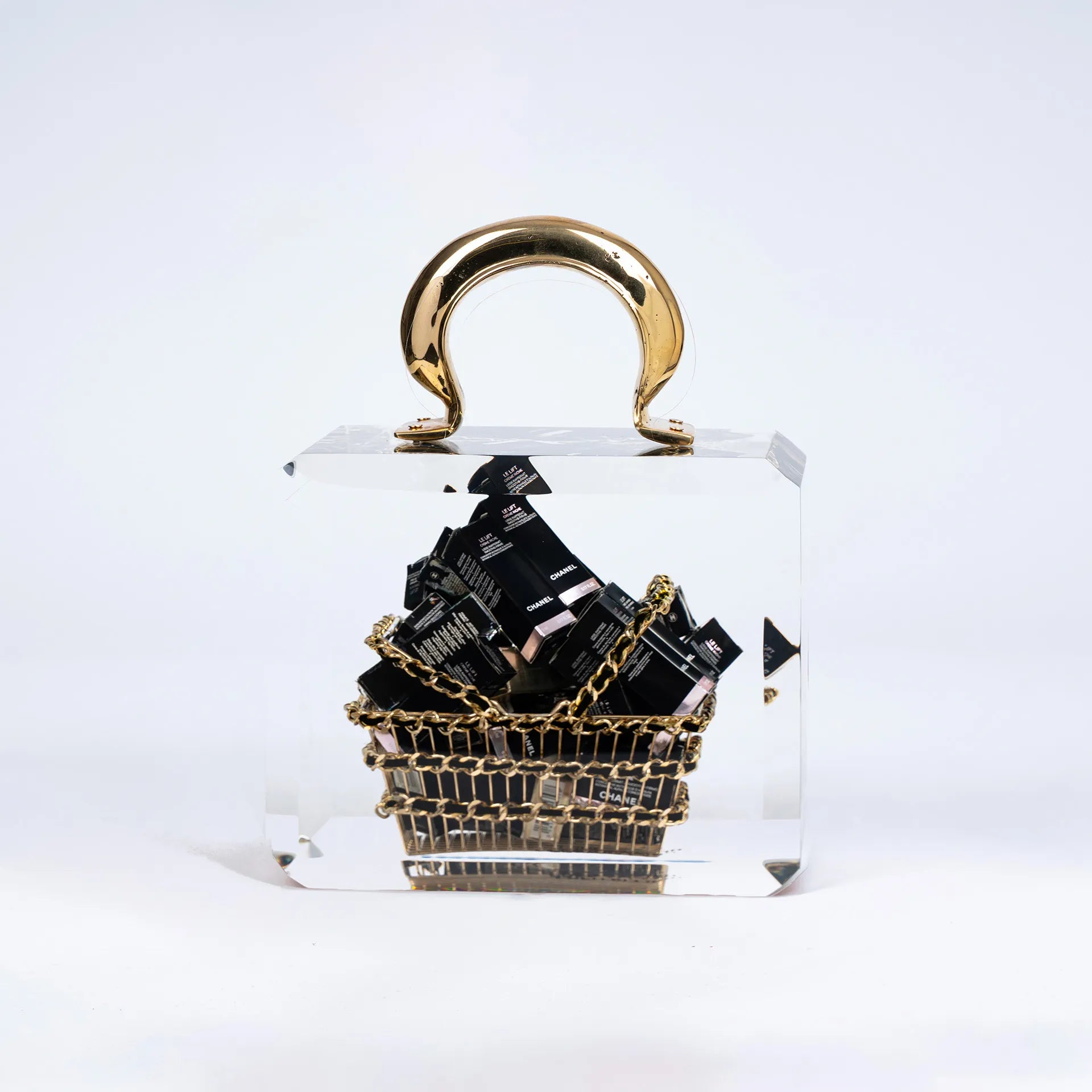 Le Chanel Overdose de Fred Allard est un luxueux basket bag transparent avec une anse dorée, contenant un petit panier doré rempli de boîtes de cosmétiques Chanel noires, le tout sur un fond blanc uni.