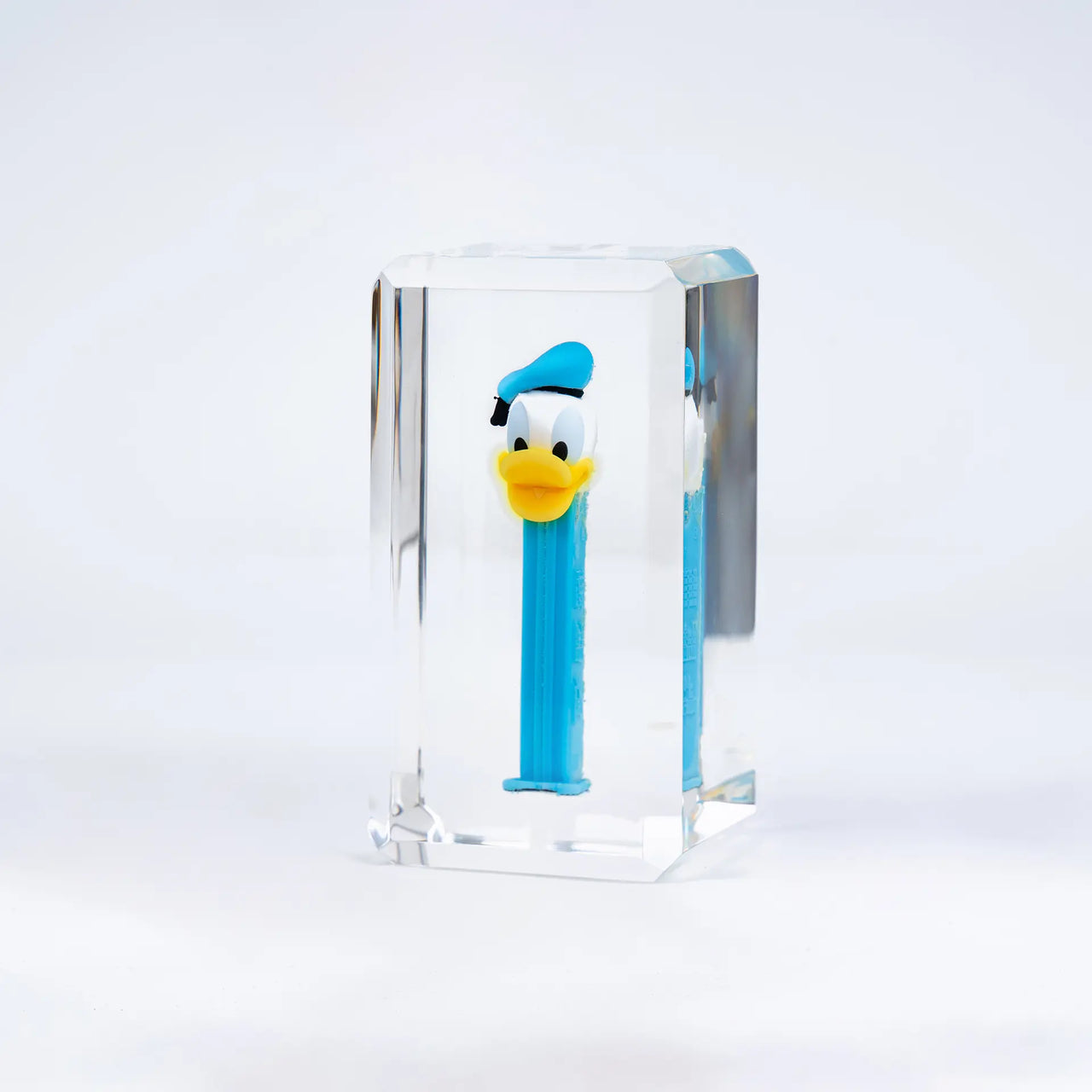 Les bonbons PEZ de Fred Allard, avec un distributeur bleu Donald Duck, sont présentés dans un bloc rectangulaire transparent sur fond blanc.