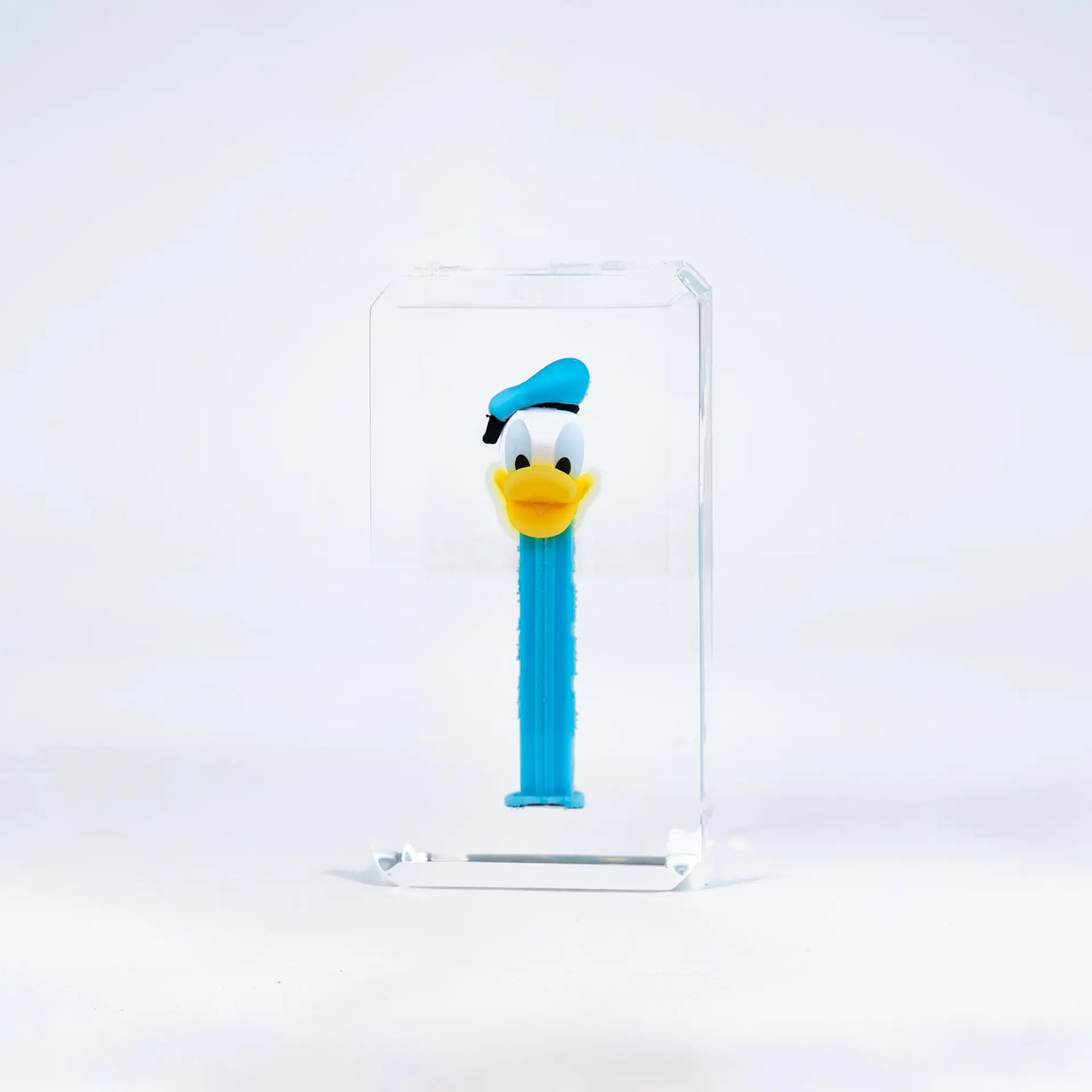 Les bonbons PEZ de Fred Allard, avec un distributeur bleu Donald Duck, sont présentés dans un bloc rectangulaire transparent sur fond blanc.