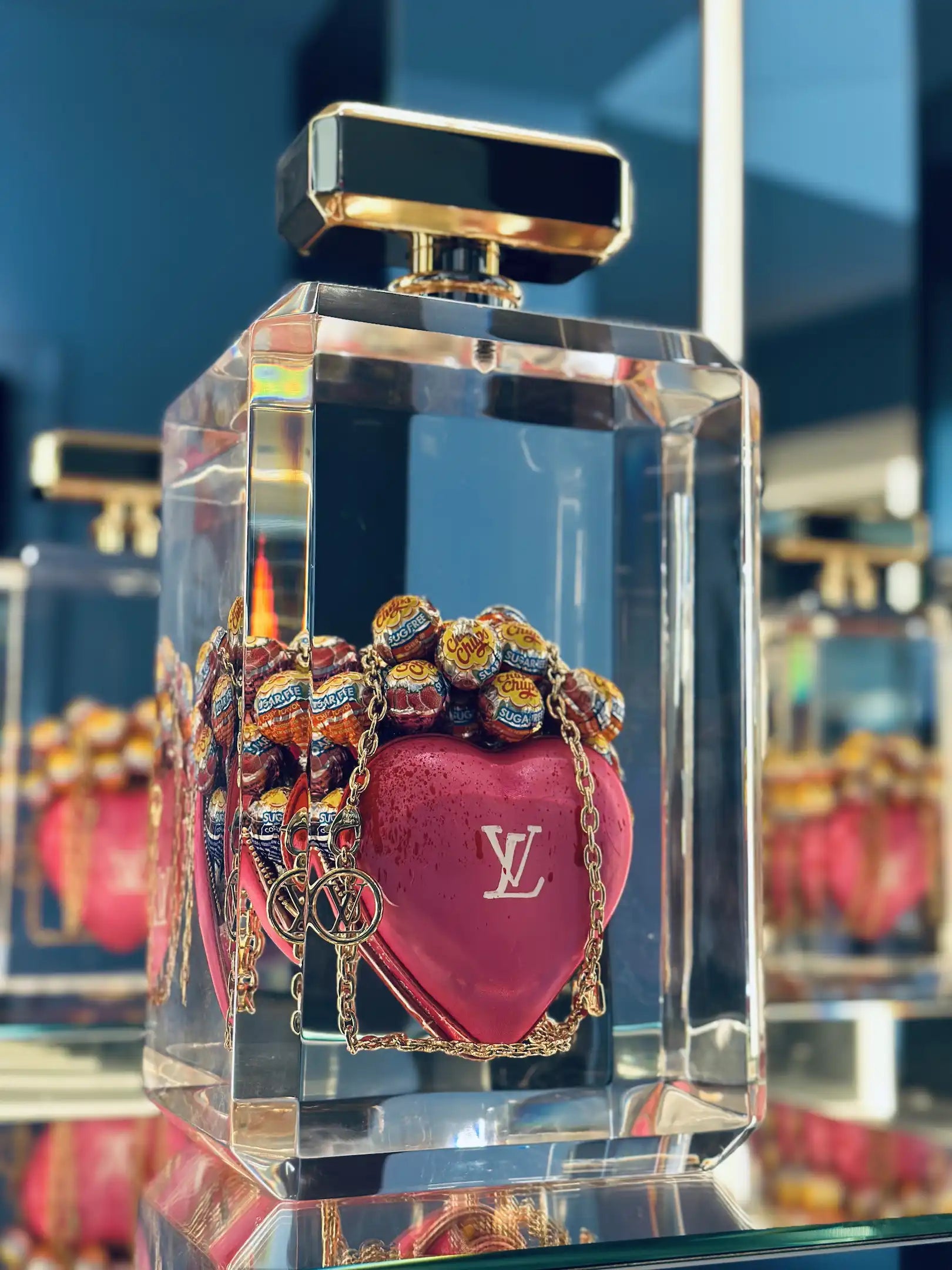 Un It bag rectangulaire transparent avec un bouchon doré contient un cœur rose étiqueté "Keep my pink heart" par Fred Allard, avec le logo Louis Vuitton sur une chaîne dorée et des sucettes Chupa Chups l'intérieur.