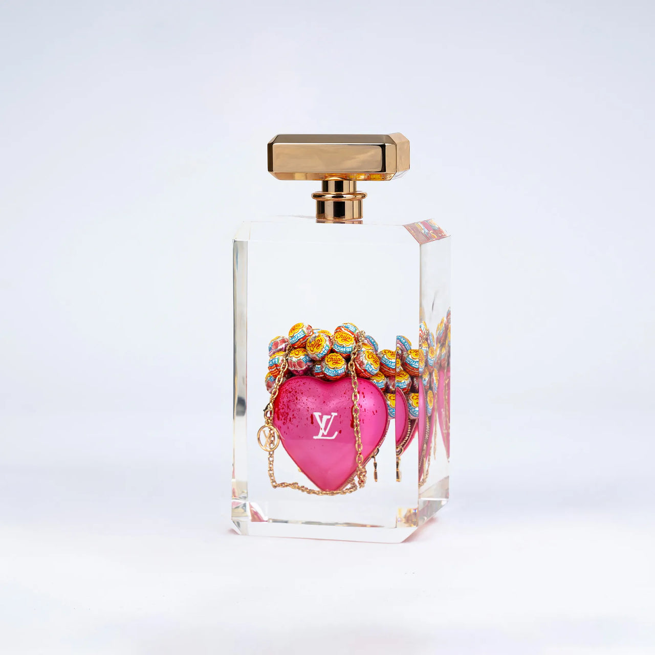 L'It bag "Keep my pink heart" de Fred Allard se présente dans un flacon rectangulaire transparent avec un bouchon doré, arborant un cœur rose et le logo Louis Vuitton, ainsi que des Chupa Chups multicolores, sur fond blanc.