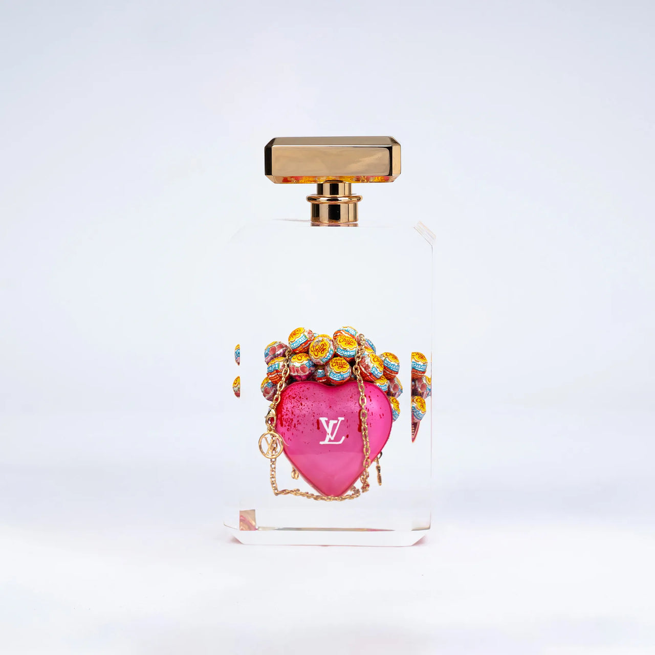 L'It bag "Keep my pink heart" de Fred Allard se présente dans un flacon rectangulaire transparent avec un bouchon doré, arborant un cœur rose et le logo Louis Vuitton, ainsi que des Chupa Chups multicolores, sur fond blanc.