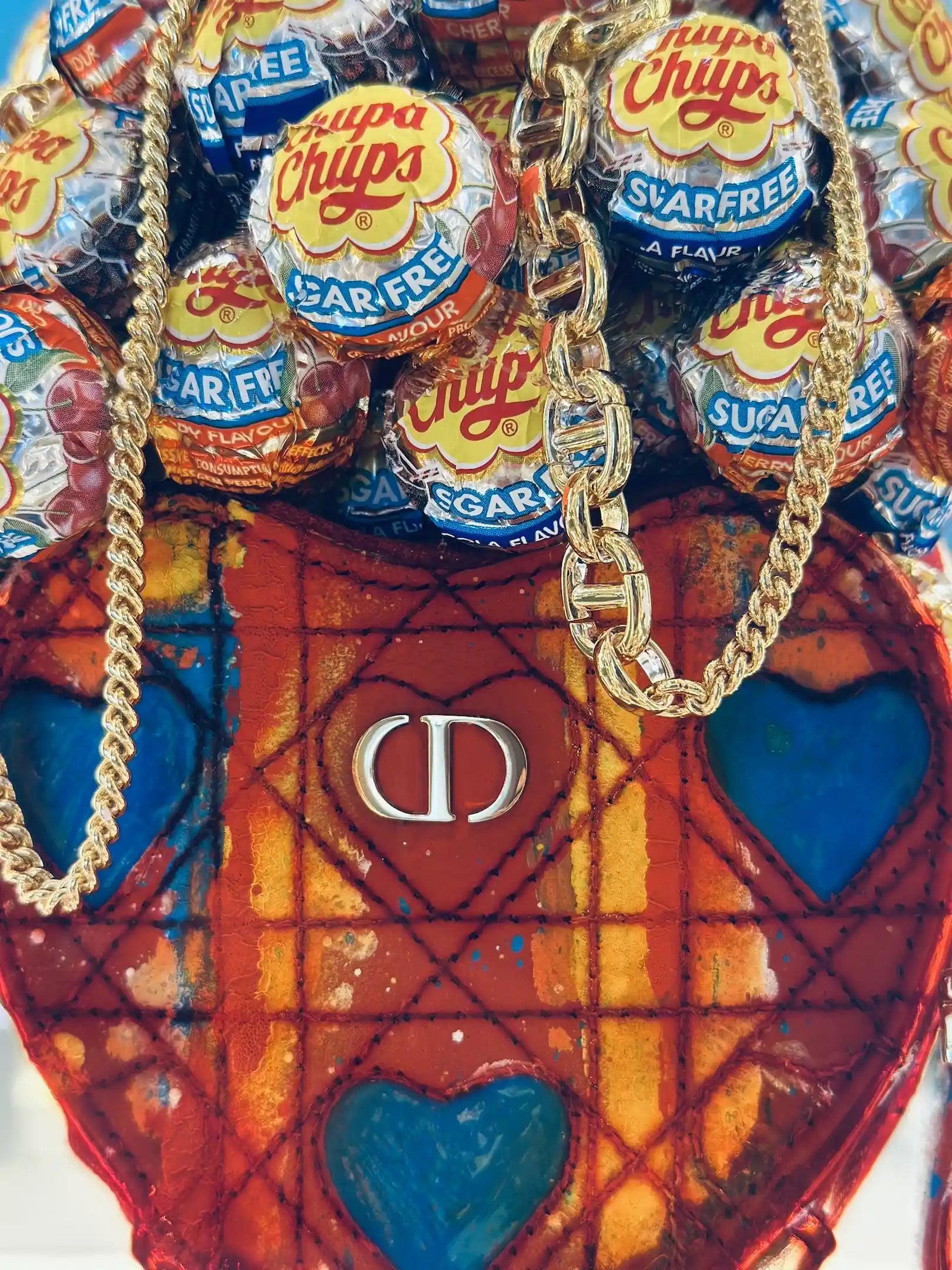 Gros plan sur le sac rouge en forme de cœur Secrètement toi de Fred Allard, orné de cœurs bleus, d'un logo Dior argenté, de chaînes dorées et de sucettes Chupa Chups.