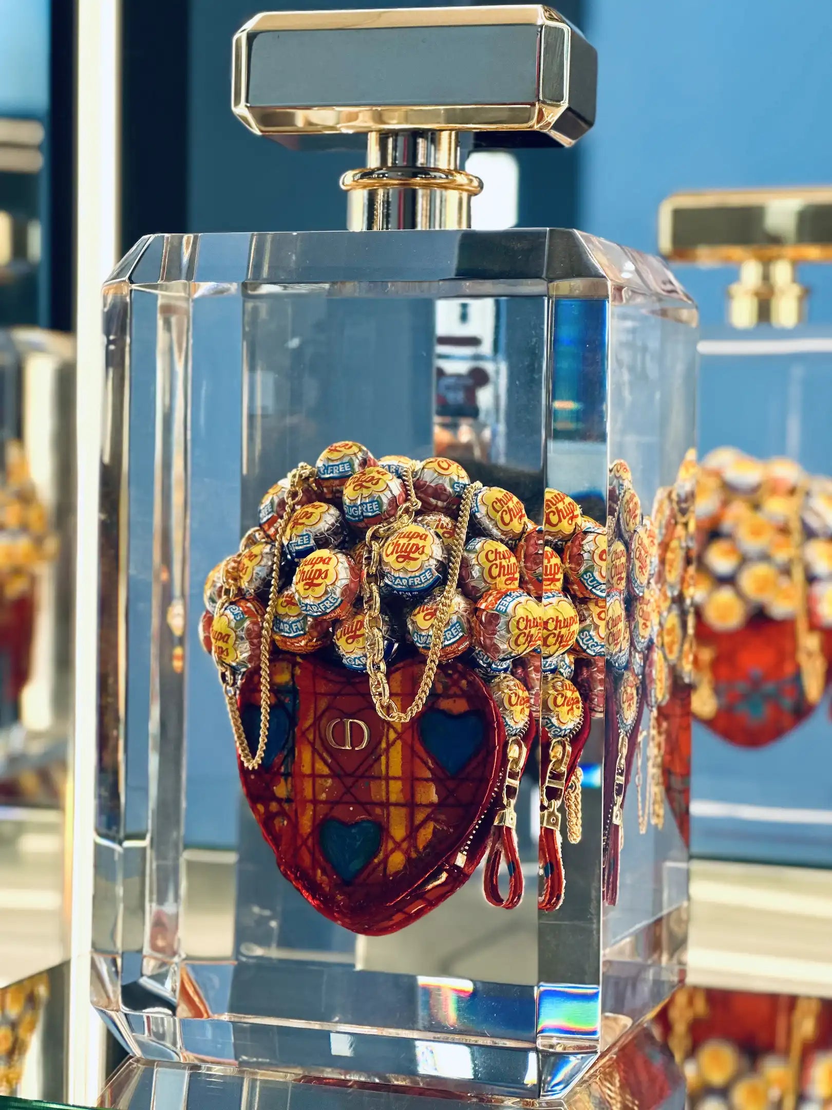 Un flacon de parfum transparent, Secrètement toi by Fred Allard, présente un cœur rouge à l'intérieur et des sucettes Chupa Chups, sur une surface réfléchissante, mettant en valeur le style artistique emblématique de Fred Allard.