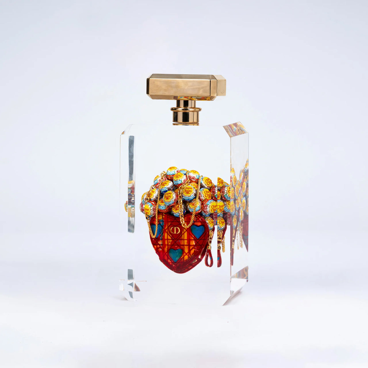 Secrètement toi de Fred Allard présente un flacon avec un bouchon rectangulaire doré et un cœur Dior orné de Chupa Chups, sur un fond blanc uni.