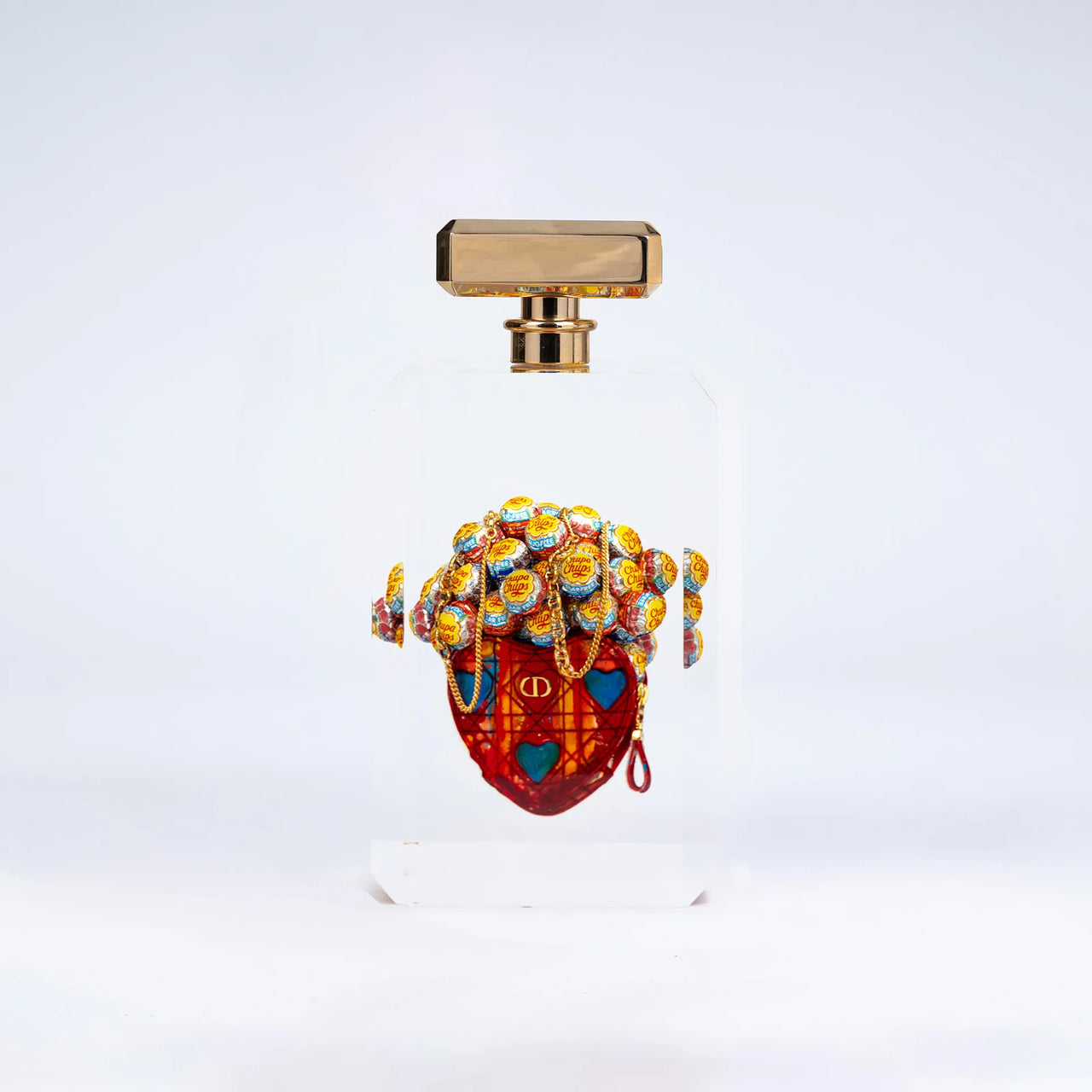 Secrètement toi de Fred Allard présente un flacon avec un bouchon rectangulaire doré et un cœur Dior orné de Chupa Chups, sur un fond blanc uni.