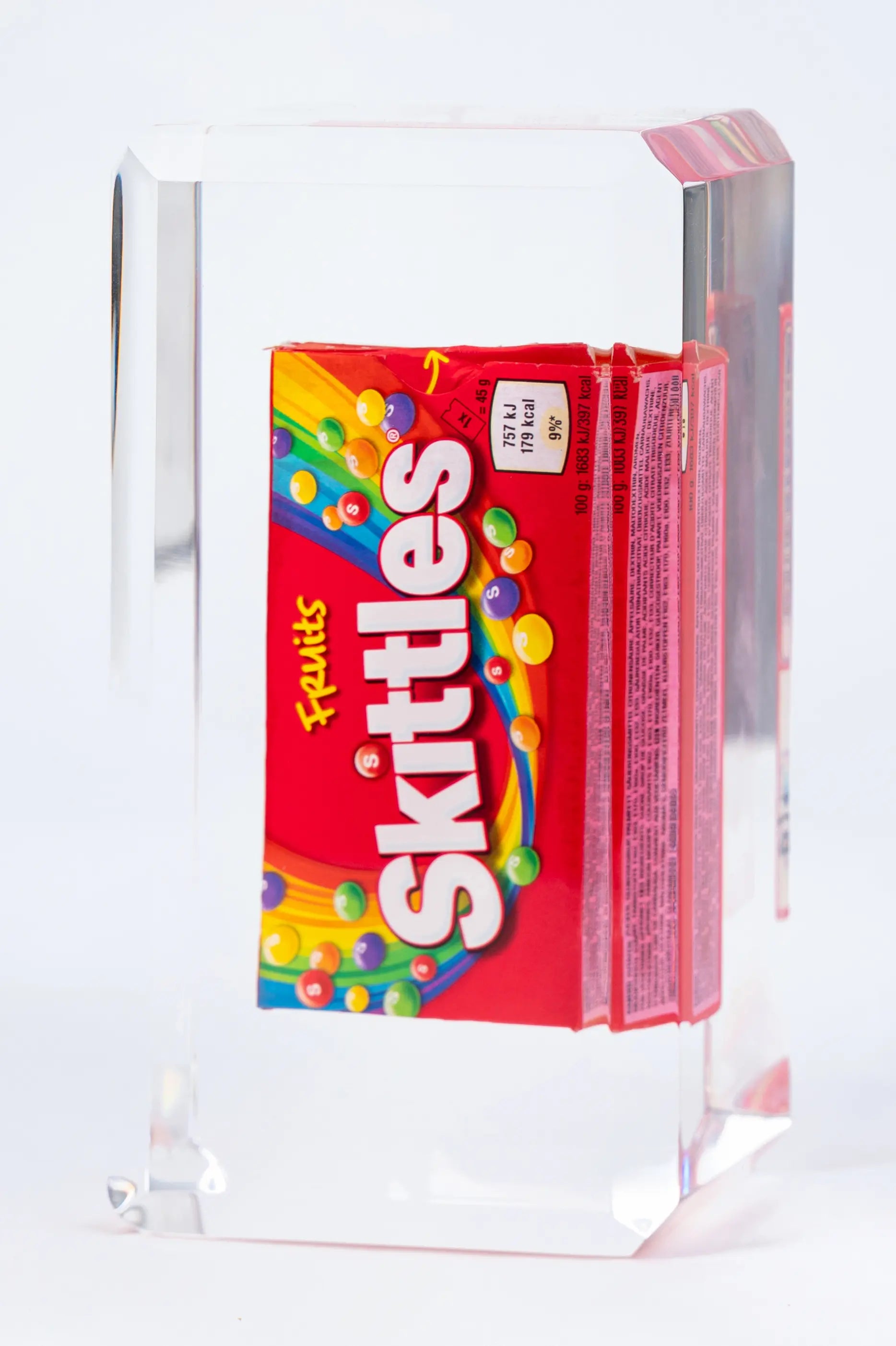Une boîte de Skittles de Fred Allard est enfermée dans un bloc rectangulaire transparent et présentée à la verticale sur un fond blanc.