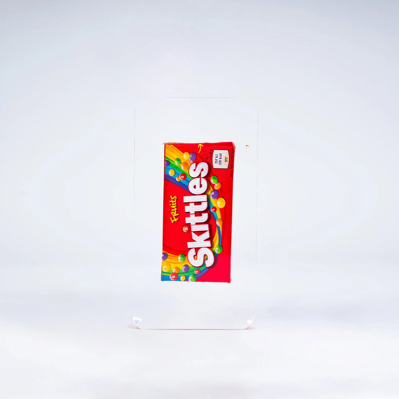 Une boîte de bonbons Skittles de Fred Allard est enfermée dans un bloc transparent et présentée sur un fond blanc.