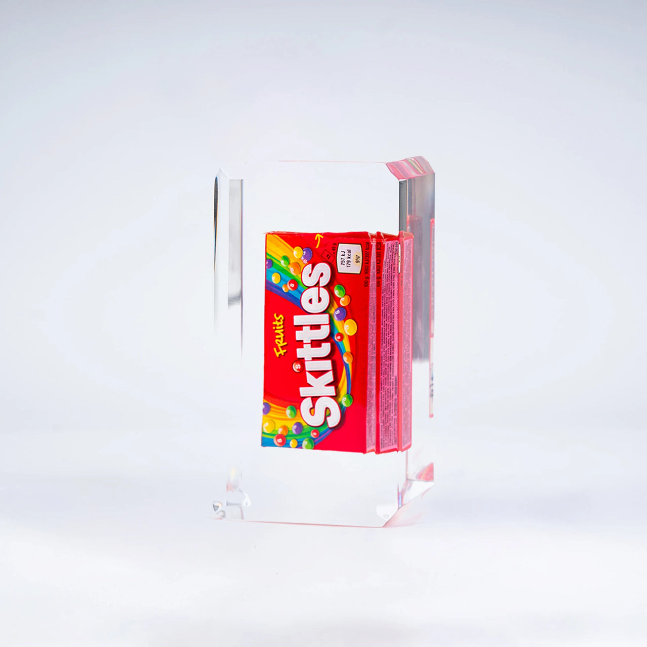 Une boîte de bonbons Skittles de Fred Allard est enfermée dans un bloc transparent et présentée sur un fond blanc.