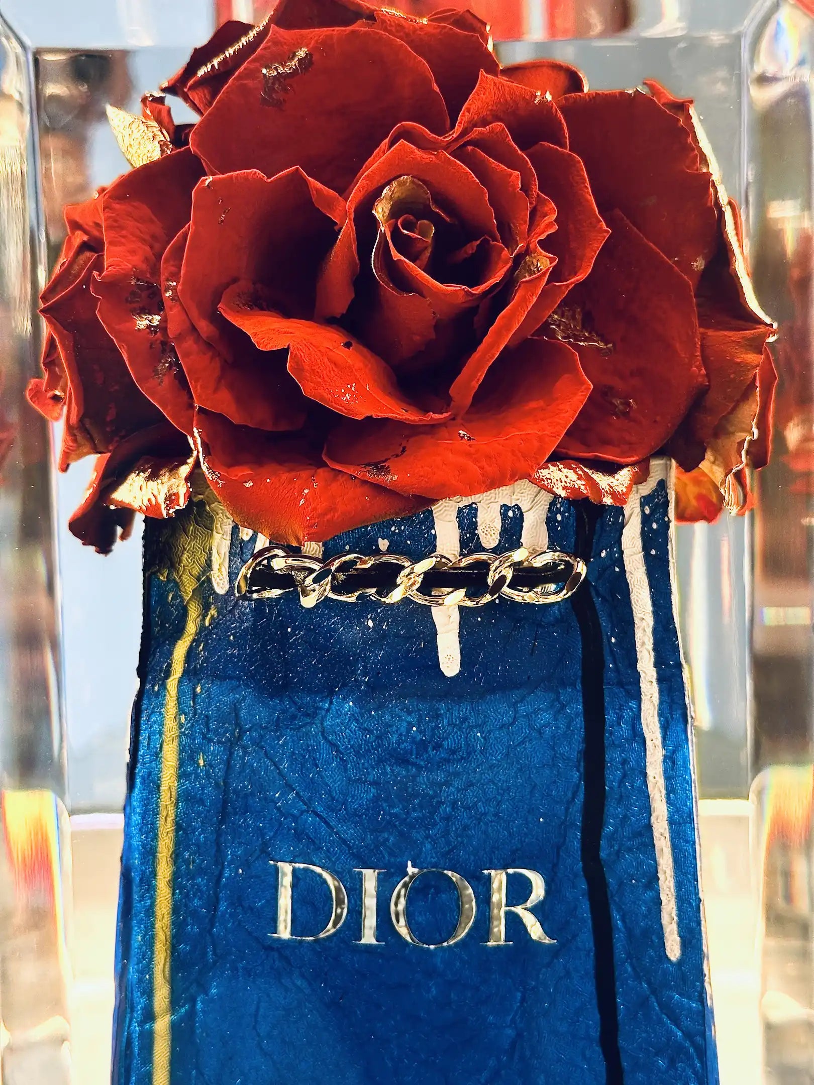 Gros plan sur True romance de Fred Allard - une composition de roses rouges dans un sac en tissu bleu portant le logo DIOR, des coulures de peinture blanches et dorées, et une chaîne argentée.