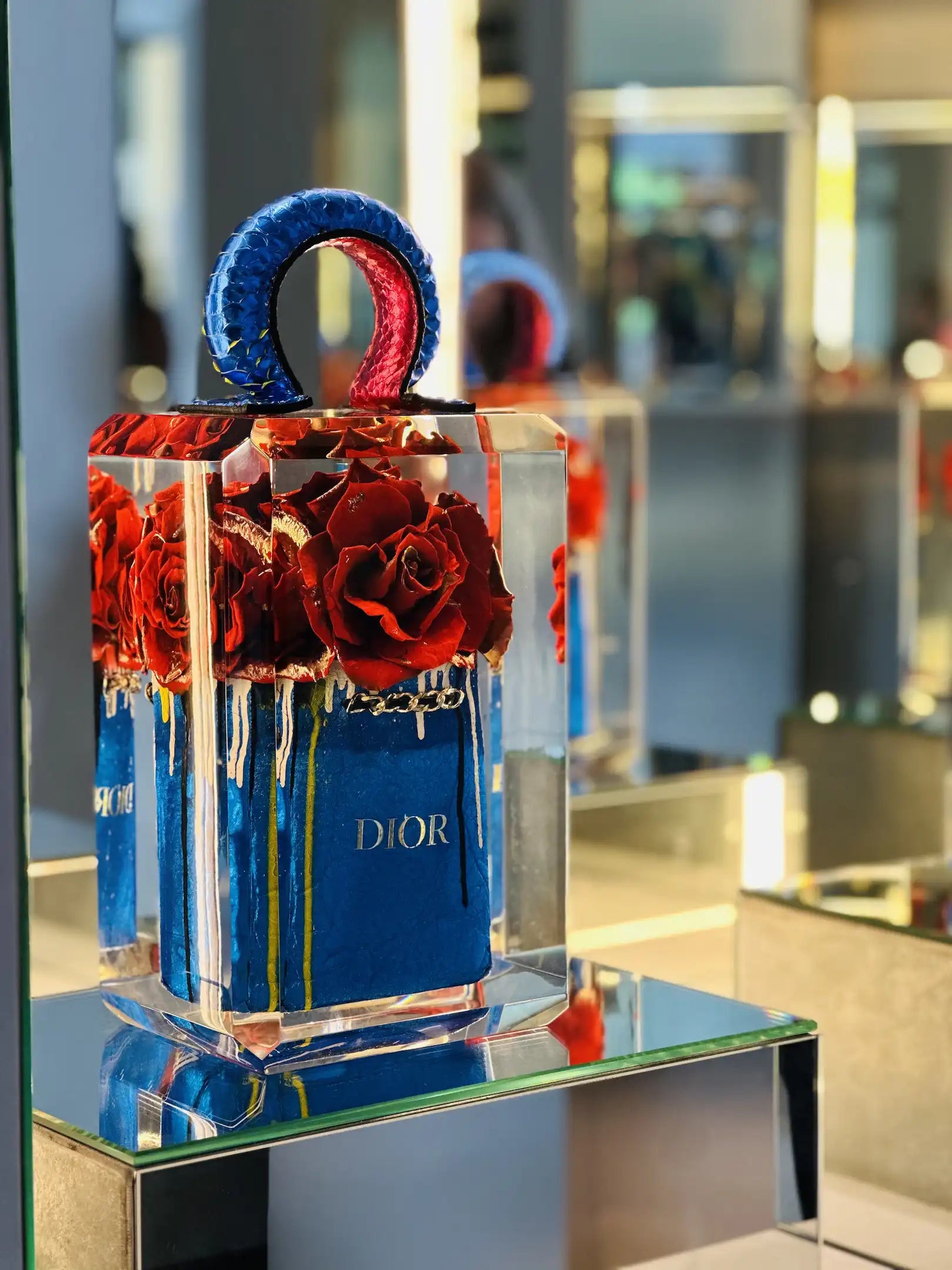 True Romance de Fred Allard présente des roses rouges dans un sac Dior bleu avec une poignée texturée bleue et rouge, enveloppée dans un bloc transparent et présentée sur une surface en miroir sous des lumières vives.