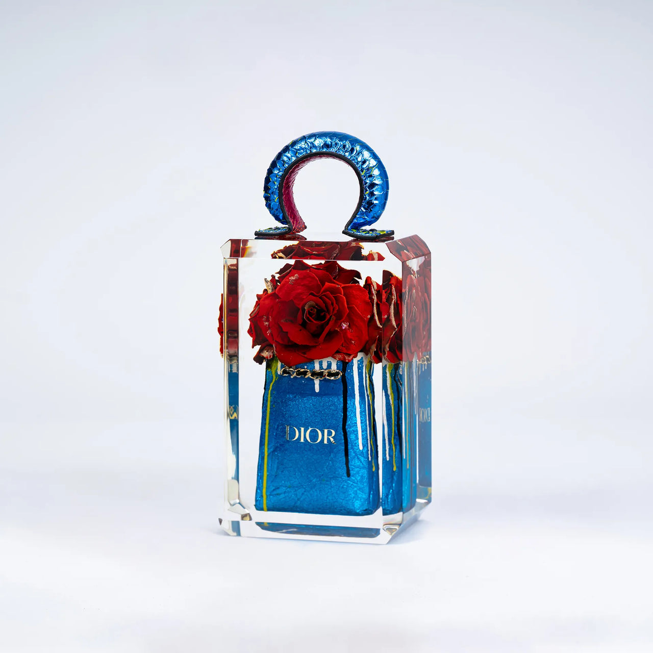 Un flacon de parfum True Romance de Fred Allard avec une surface réfléchissante présente une grappe de roses rouges à l'intérieur et un sac Dior, le tout sur un fond blanc uni.