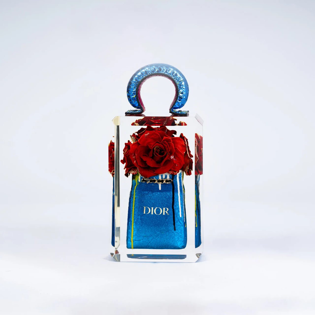 Un flacon de parfum True Romance de Fred Allard avec une surface réfléchissante présente une grappe de roses rouges à l'intérieur et un sac Dior, le tout sur un fond blanc uni.