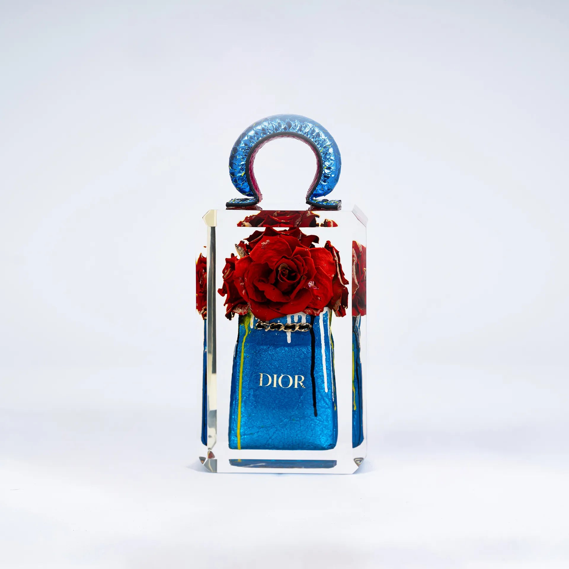 Un flacon de parfum True Romance de Fred Allard avec une surface réfléchissante présente une grappe de roses rouges à l'intérieur et un sac Dior, le tout sur un fond blanc uni.