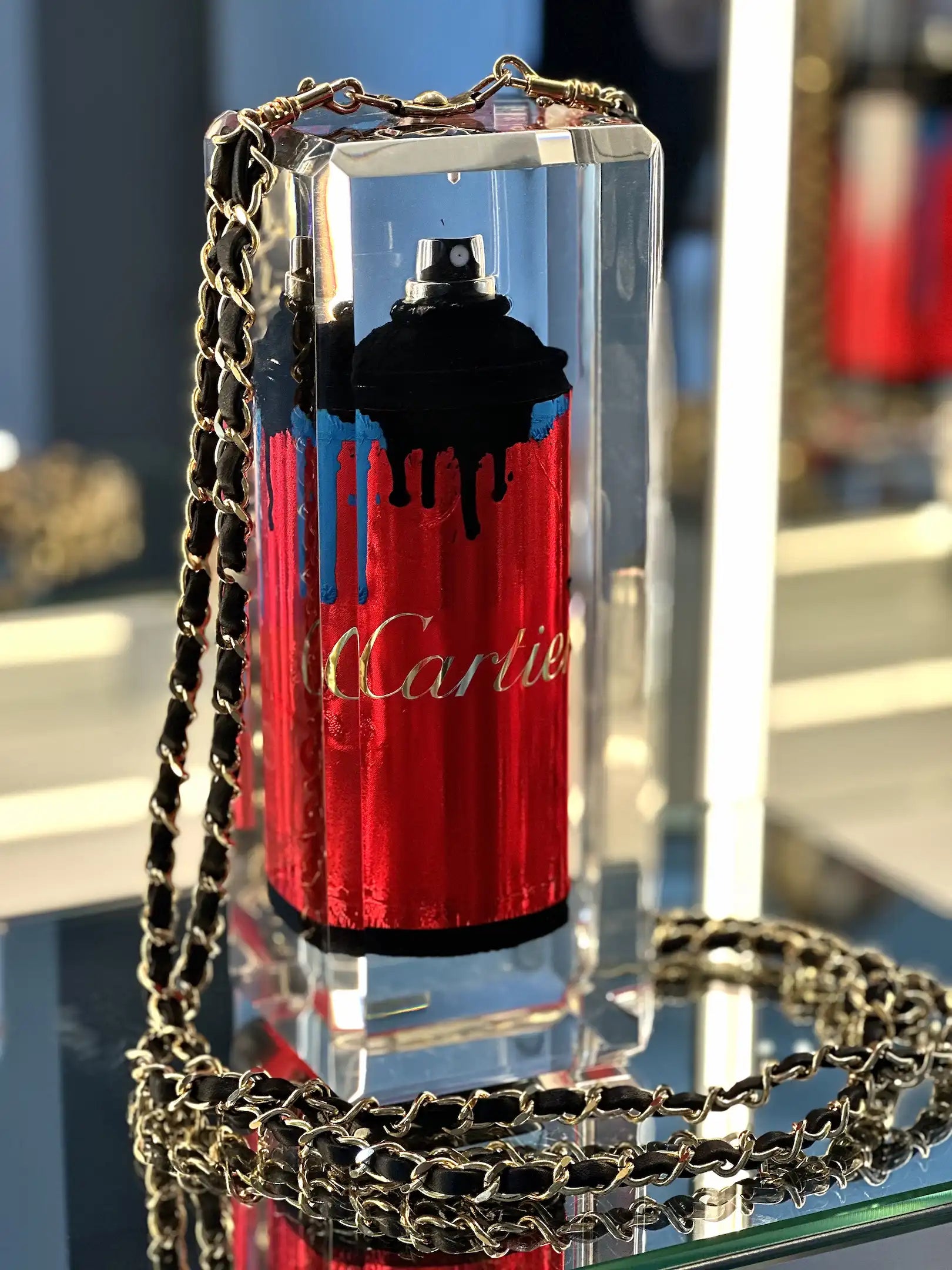 Undercover by Fred Allard est une oeuvre transparente avec, à l'intérieur, une bombe de peinture rouge "Cartier", avec des gouttes noires et une bande bleue. Il est muni d'une chaîne en or et est présenté sur une surface réfléchissante.