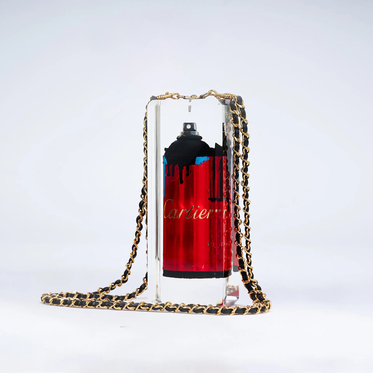Une bombe de peinture rouge étiquetée "Undercover" par Fred Allard, avec des gouttes noires, est enfermée dans un bloc rectangulaire transparent et ornée d'une chaîne dorée et noire, sur un fond blanc uni.