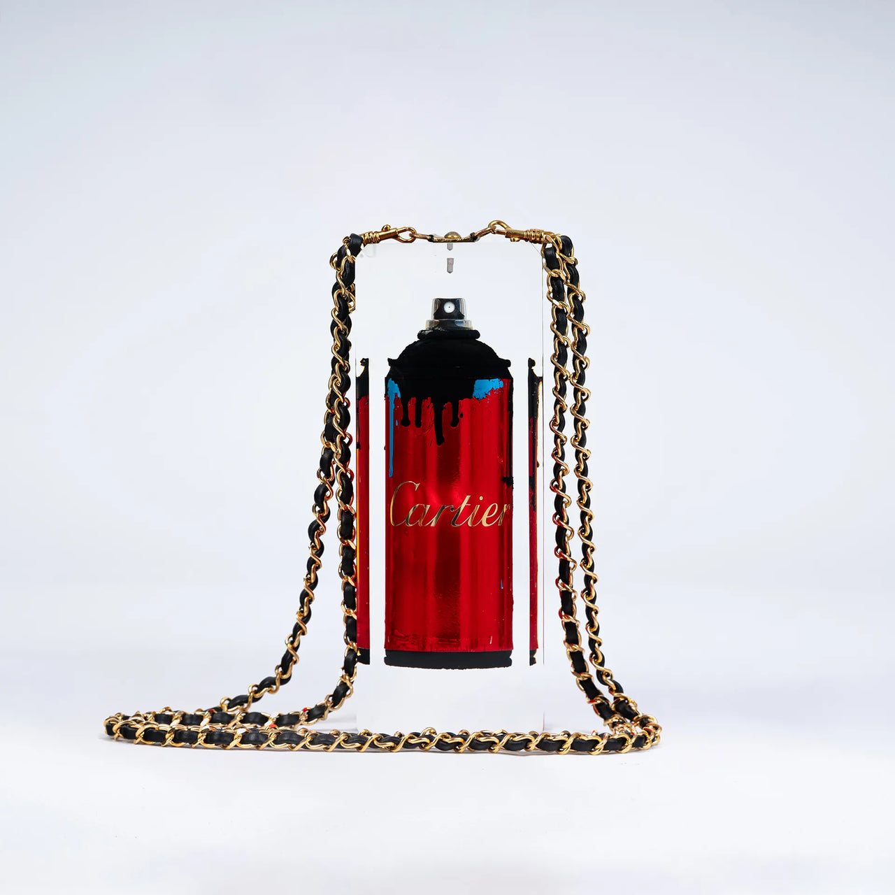 Une bombe de peinture rouge étiquetée "Undercover" par Fred Allard, avec des gouttes noires, est enfermée dans un bloc rectangulaire transparent et ornée d'une chaîne dorée et noire, sur un fond blanc uni.